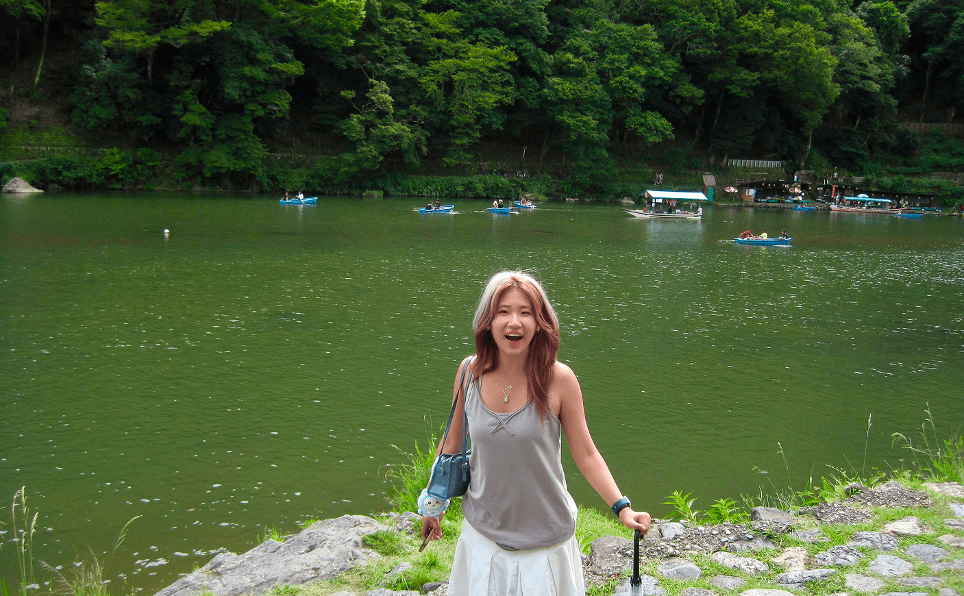 Jodie standing beside a green lake with rental paddle boats on the water and forested hills behind her.
